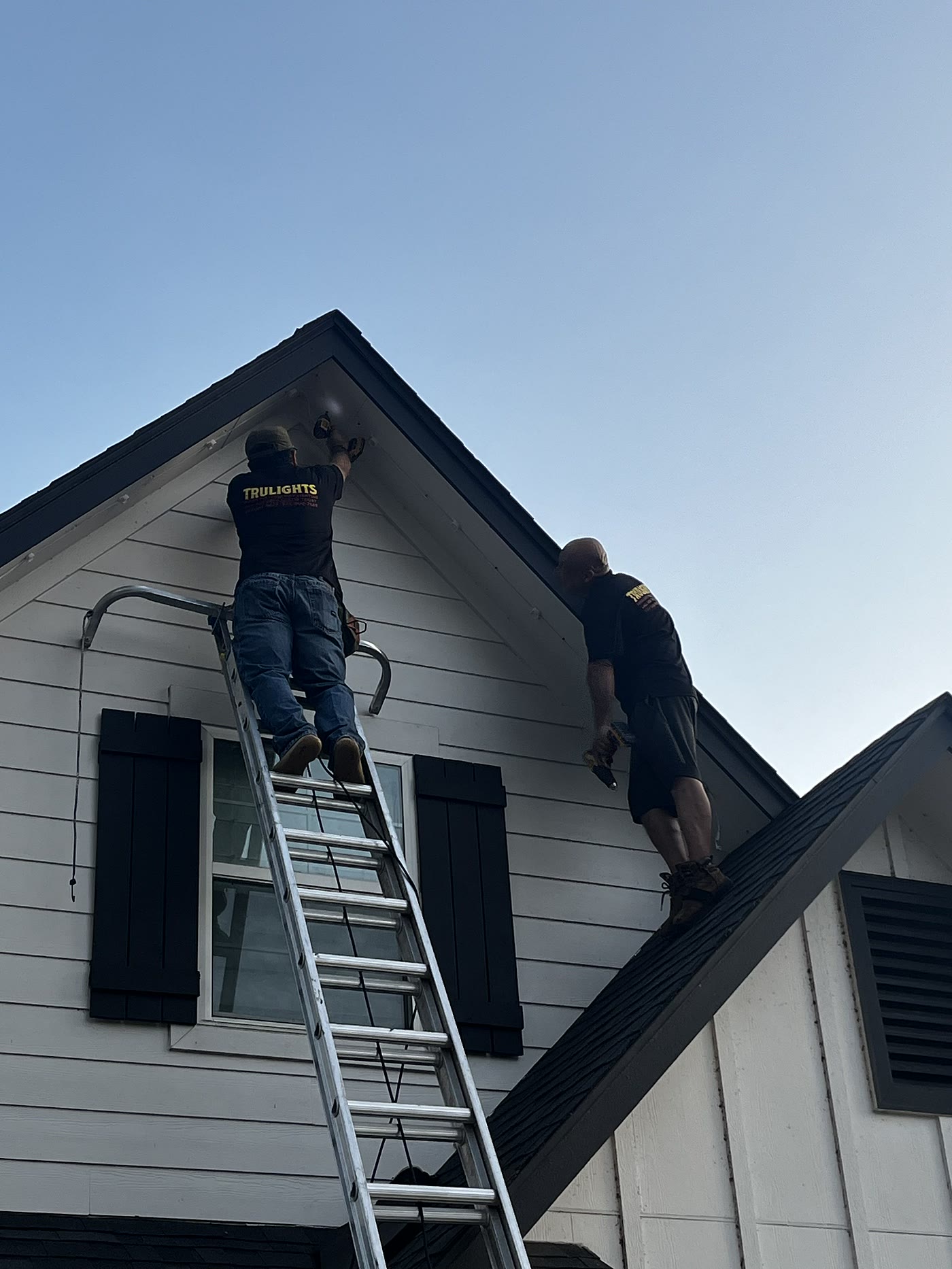 Two TruLights installers working a gable peak from a ladder and the roof